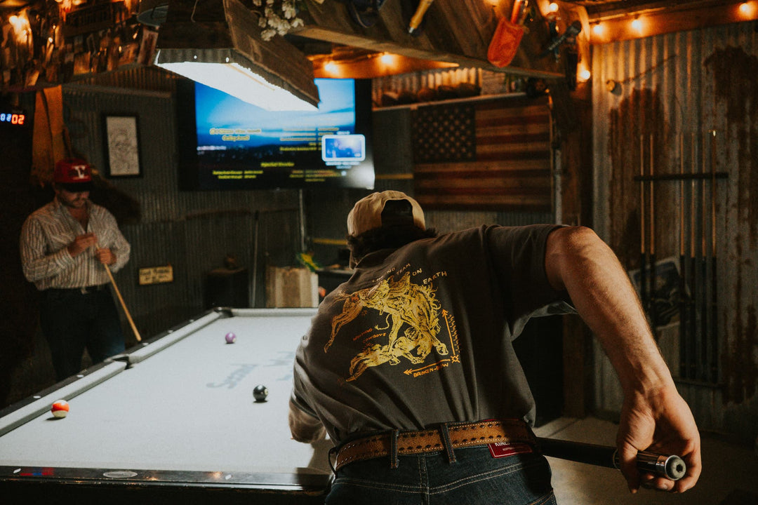 Man leaning over a pool table in a rustic bar, wearing a gray tee with a yellow hand-drawn bronco rider print on the back.